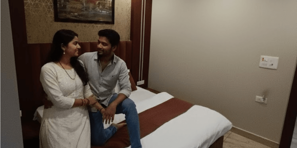 Couple relaxing in a clean and comfortable deluxe hotel room at Navodaya Sarovar, Orai, featuring modern interiors, cozy bedding, and a peaceful stay experience.
